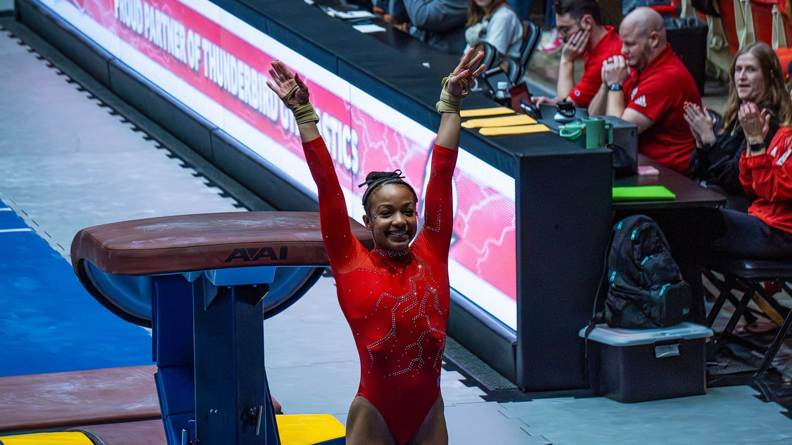 A Southern Utah gymnast in a red leotard salutes the judges with both arms raised beside the vault table during the Feb. 14, 2026 meet at Oregon State.