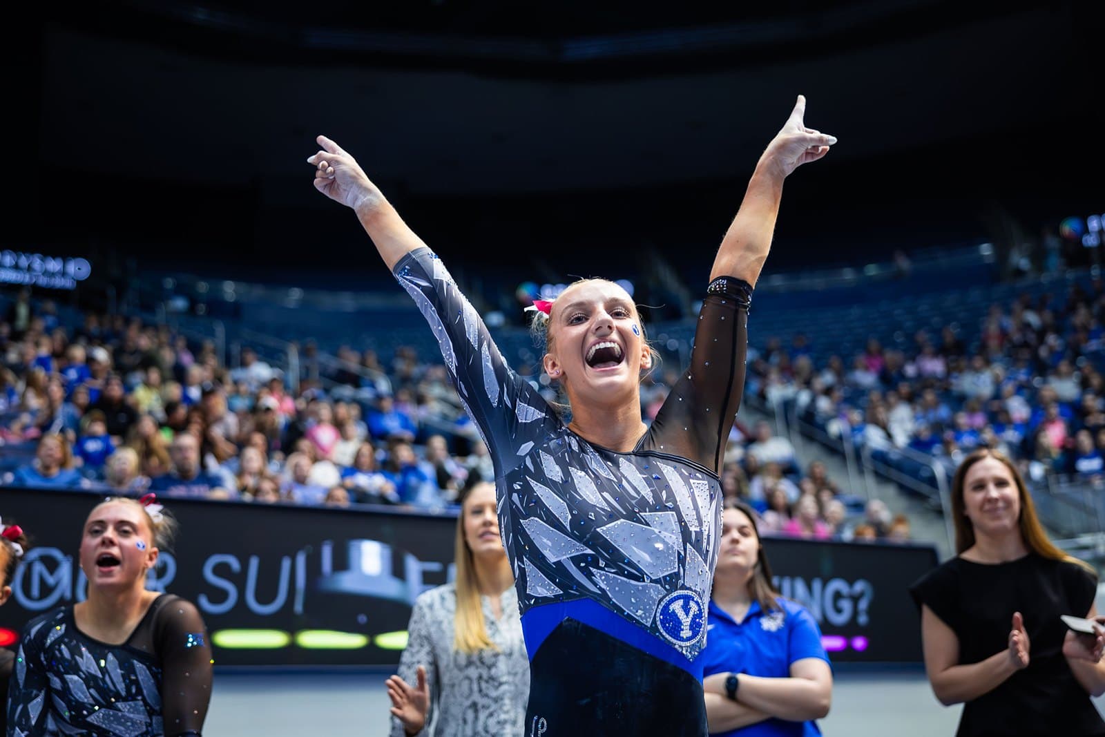 A BYU gymnast in a dark patterned leotard raises both arms and cheers at the end of a routine as teammates and coaches react behind her