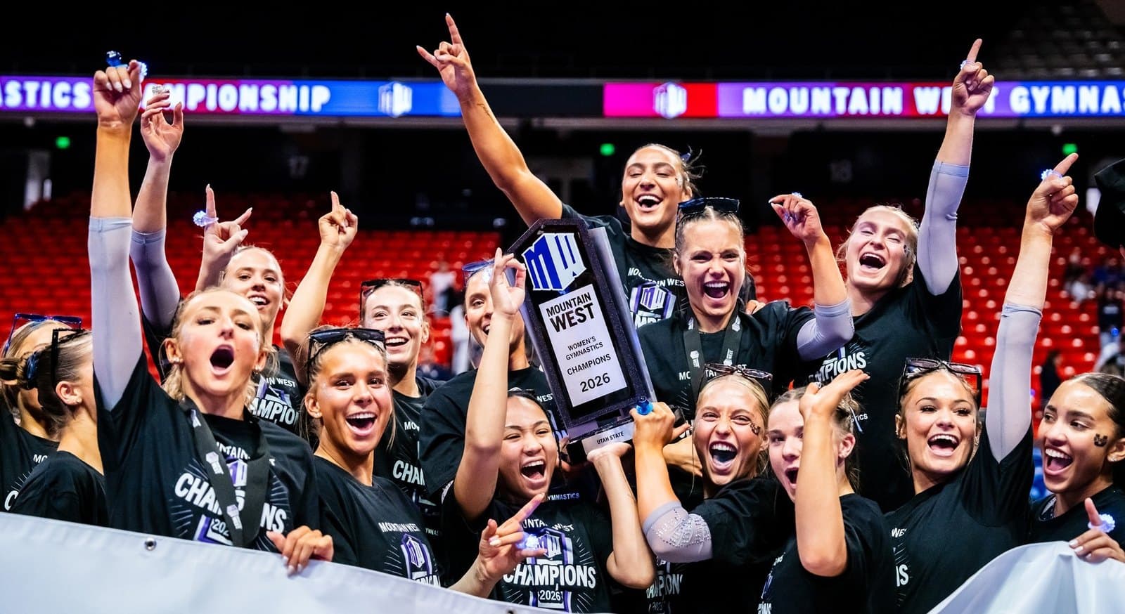Utah State gymnasts in black championship shirts raise their arms around the Mountain West trophy under the championship banner