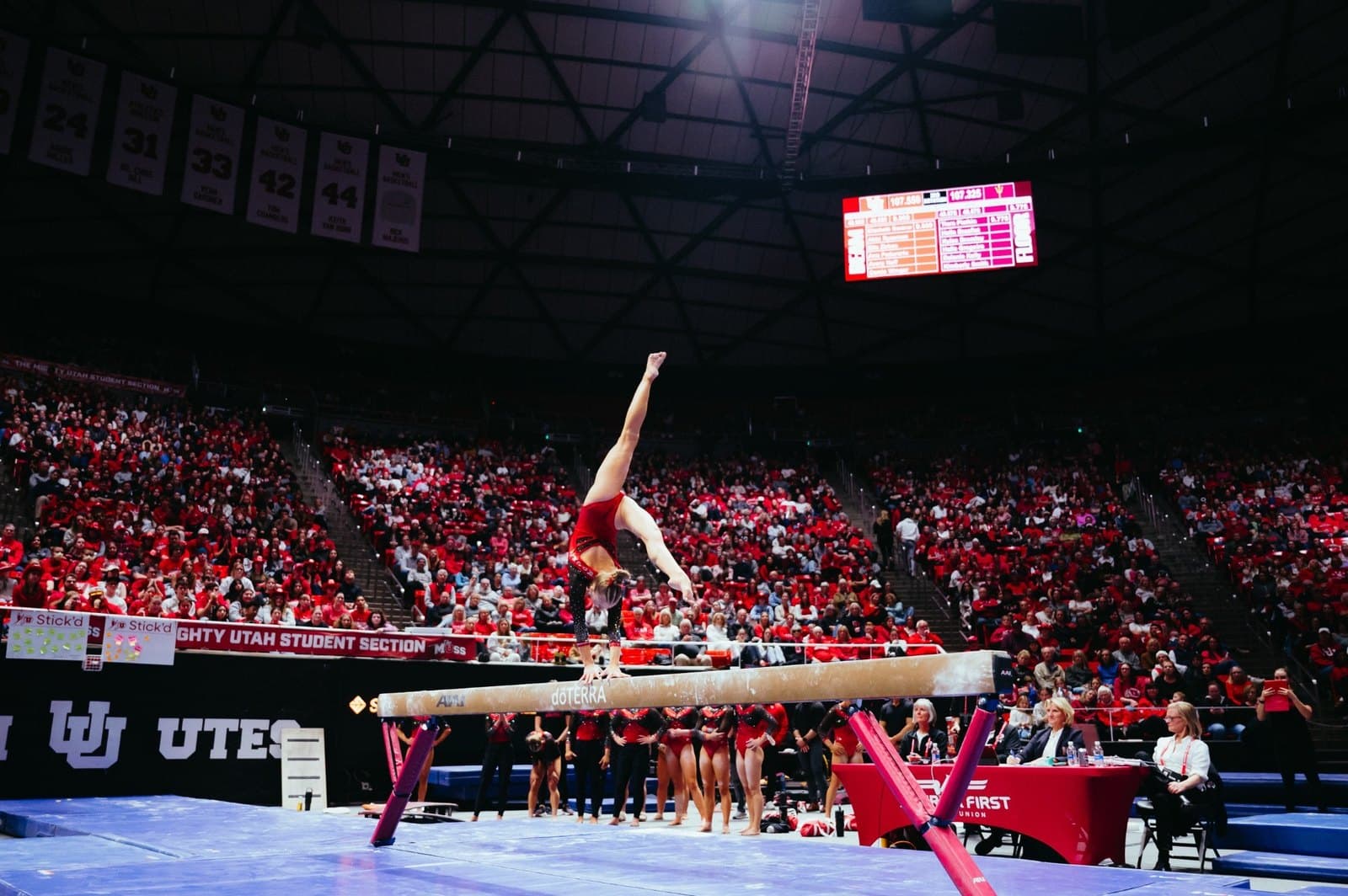 A Utah gymnast in a red leotard leaps on the balance beam in front of the crowd at the Jon M. Huntsman Center during the Jan. 30, 2026 meet against Arizona State.