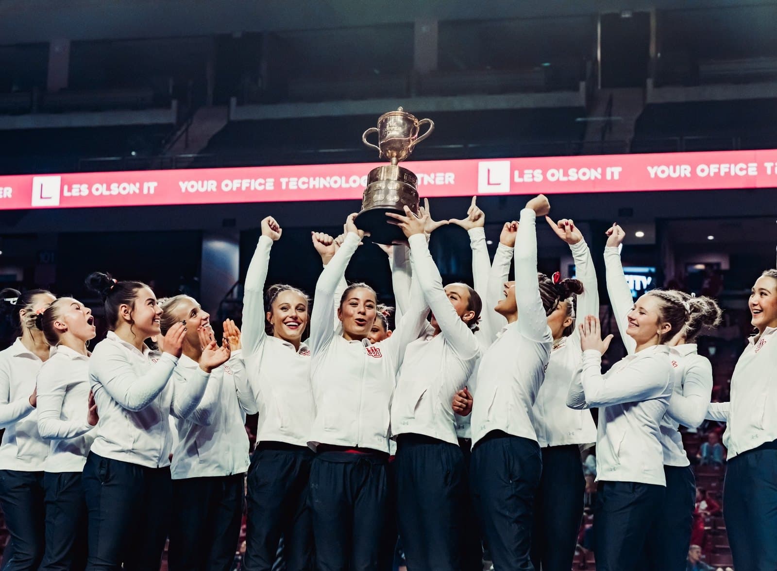 Utah gymnasts in white warm-up jackets raise the Best of Utah trophy under the Les Olson IT banner at the Maverik Center